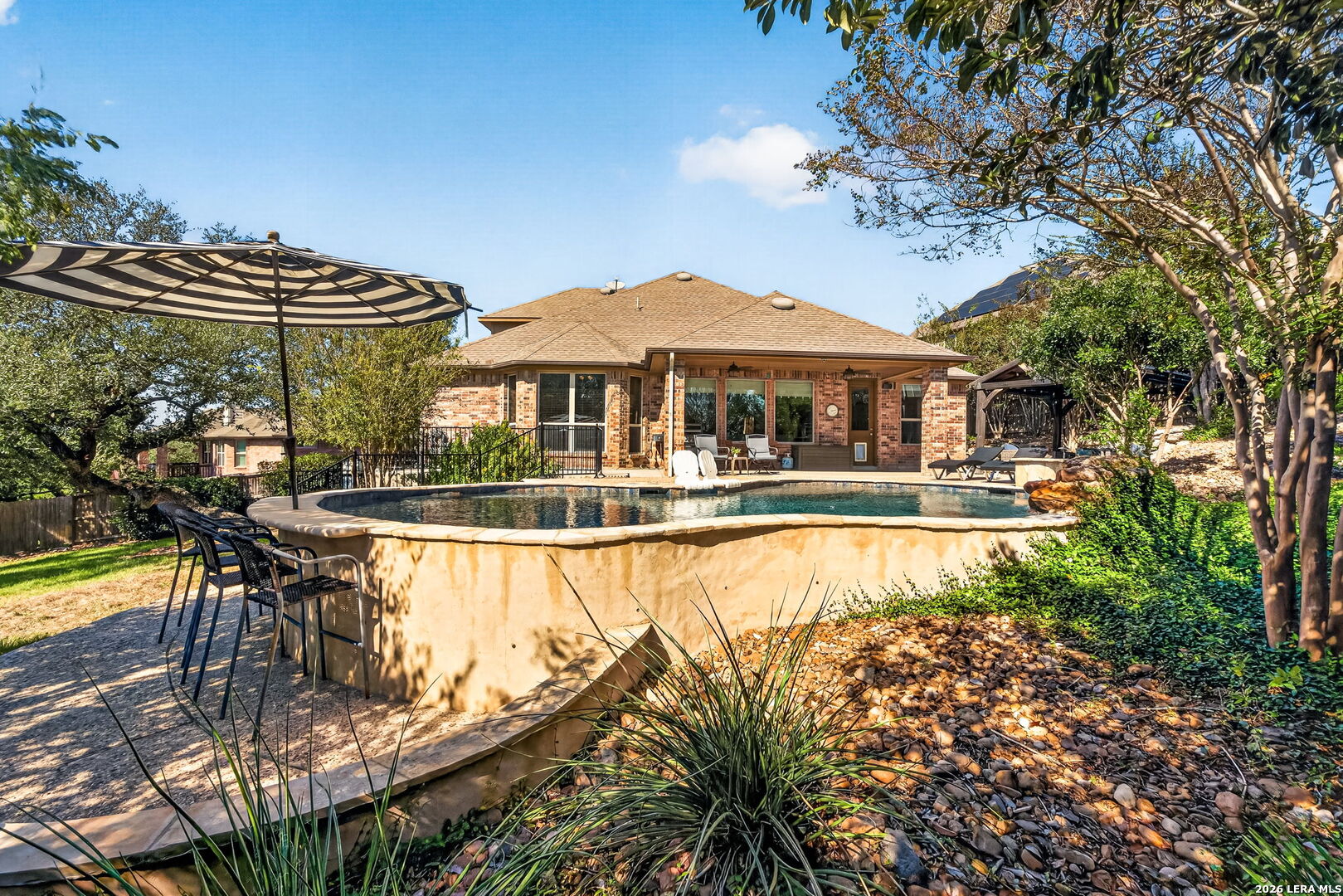 Back of a brick home overlooking a swimming pool, with a shaded patio and outdoor seating.