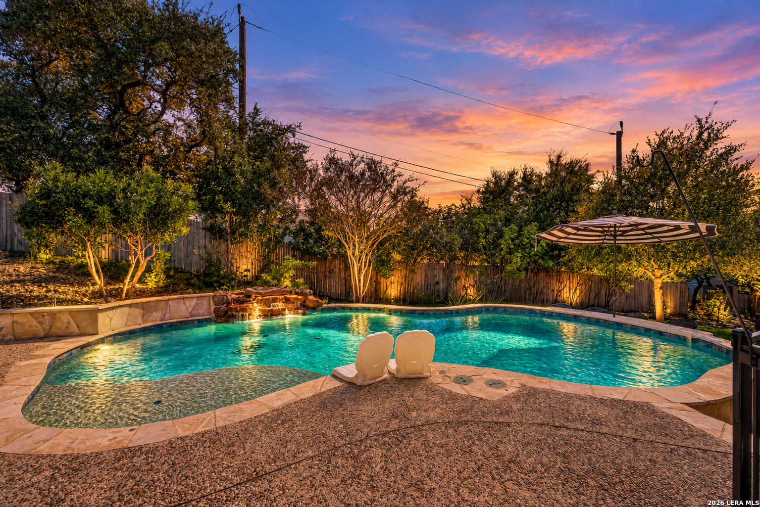 Backyard swimming pool with a sun shelf, umbrella, and illuminated landscaping at dusk.