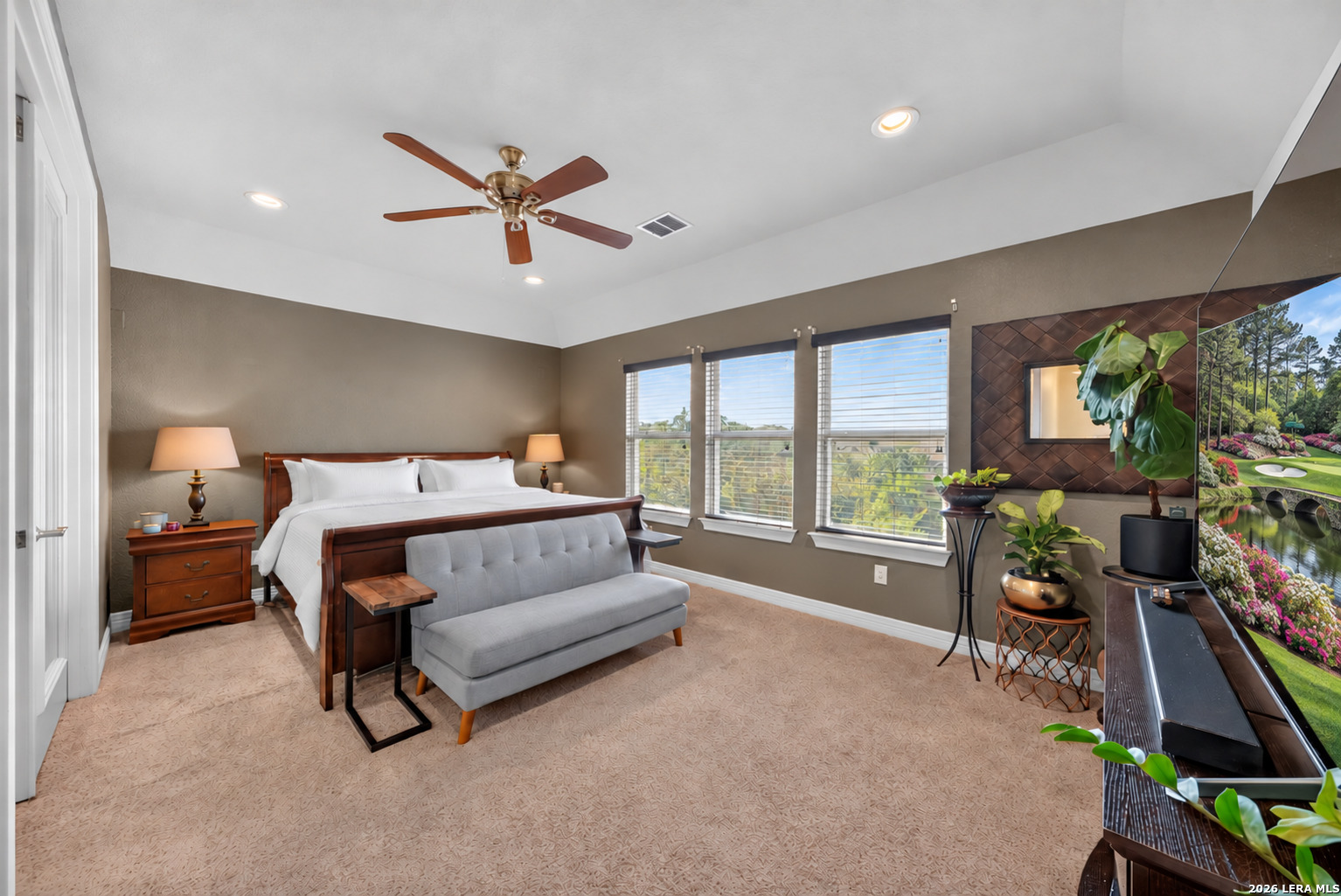 Primary bedroom with a dark wood bed, gray loveseat, and three large windows.