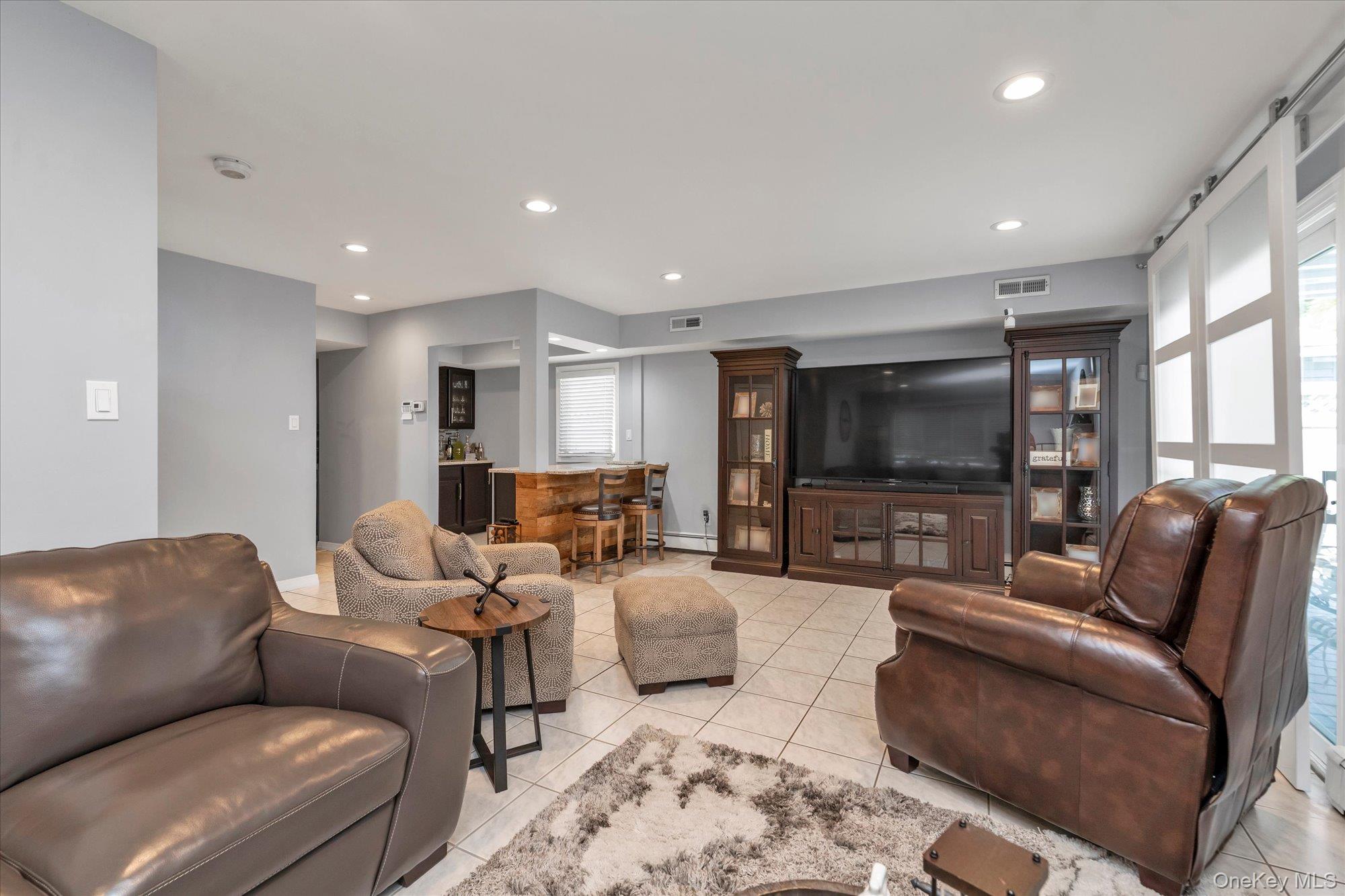 Furnished living room with brown leather seating, tiled floors, and a large television between dark wood cabinets.