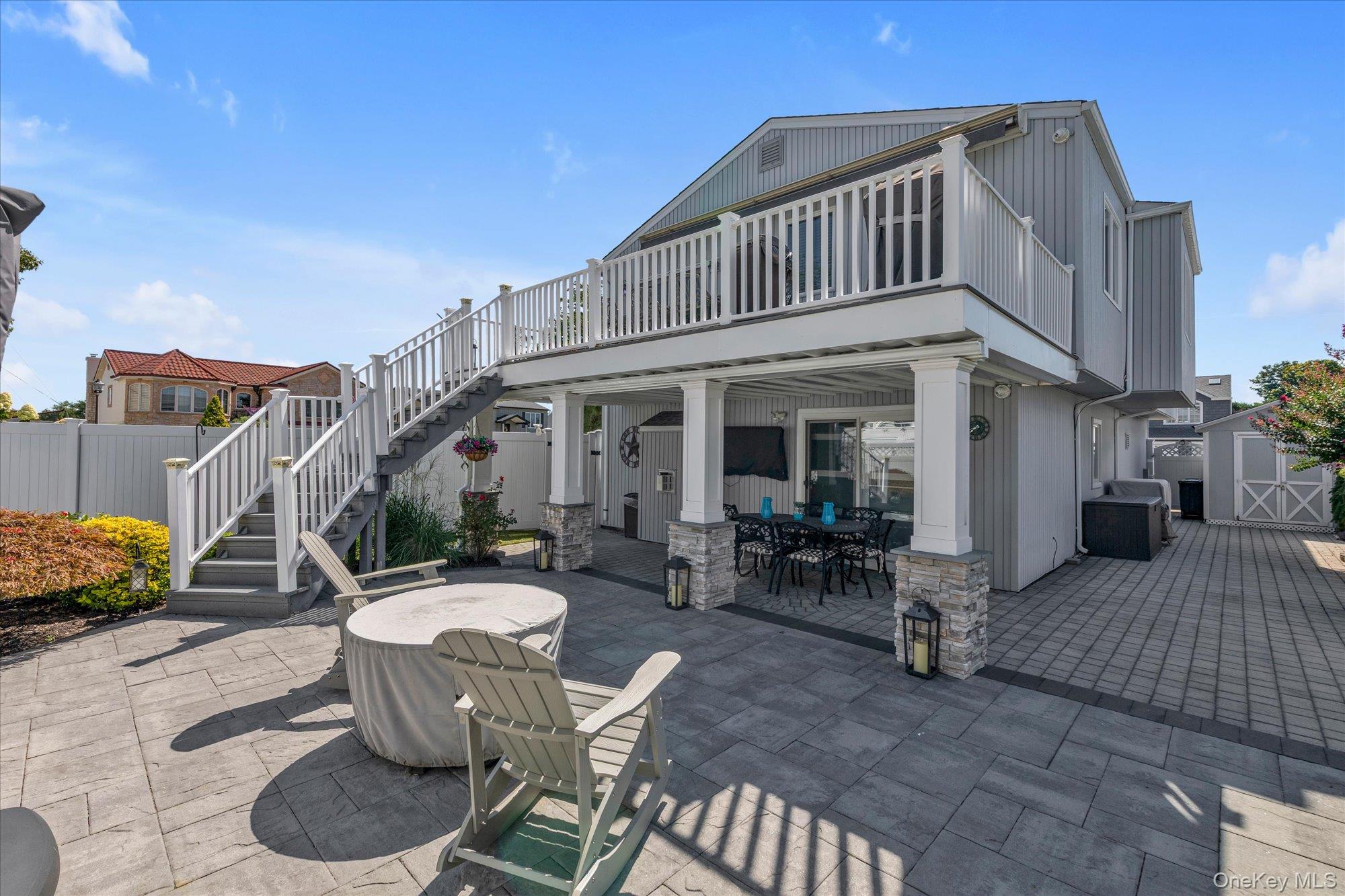 Raised gray home with a white-railed deck above a spacious furnished paved patio.