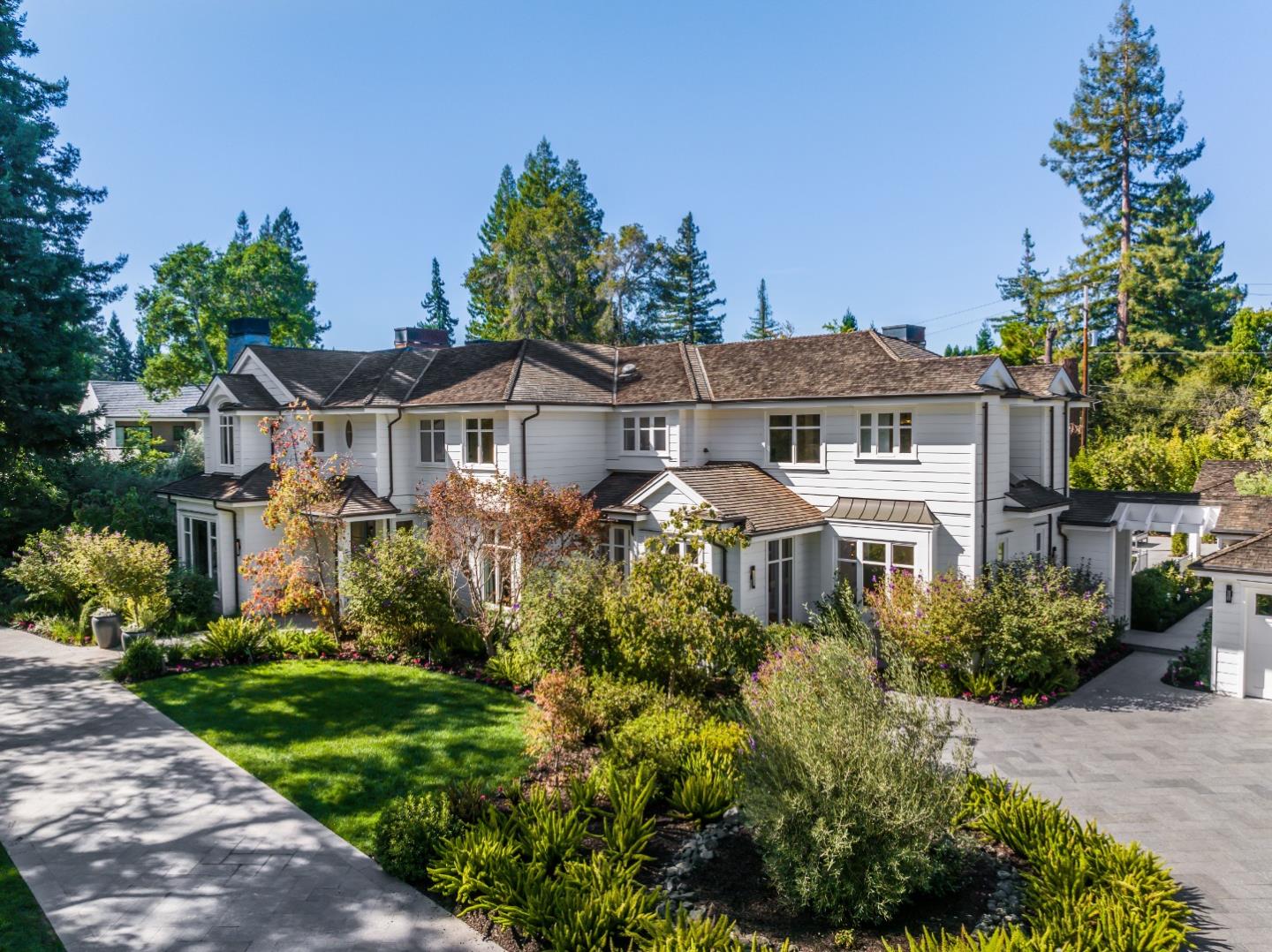 White multi-story house surrounded by landscaped gardens and tall evergreen trees.