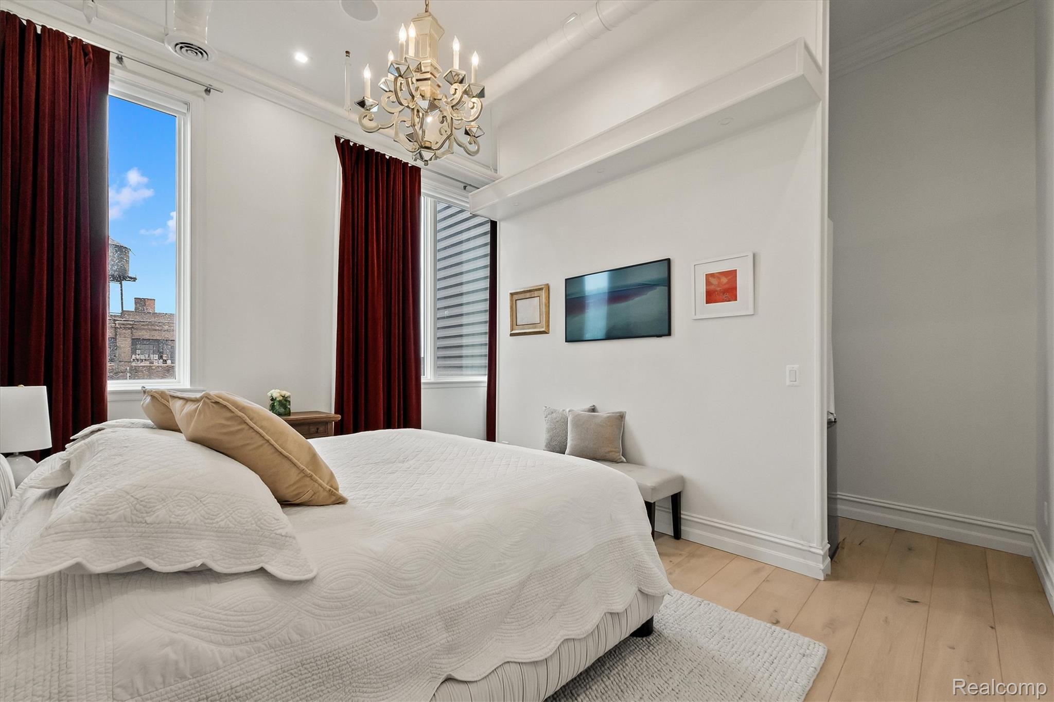 Bedroom with a white quilted bed, burgundy curtains, tall windows, and a chandelier.