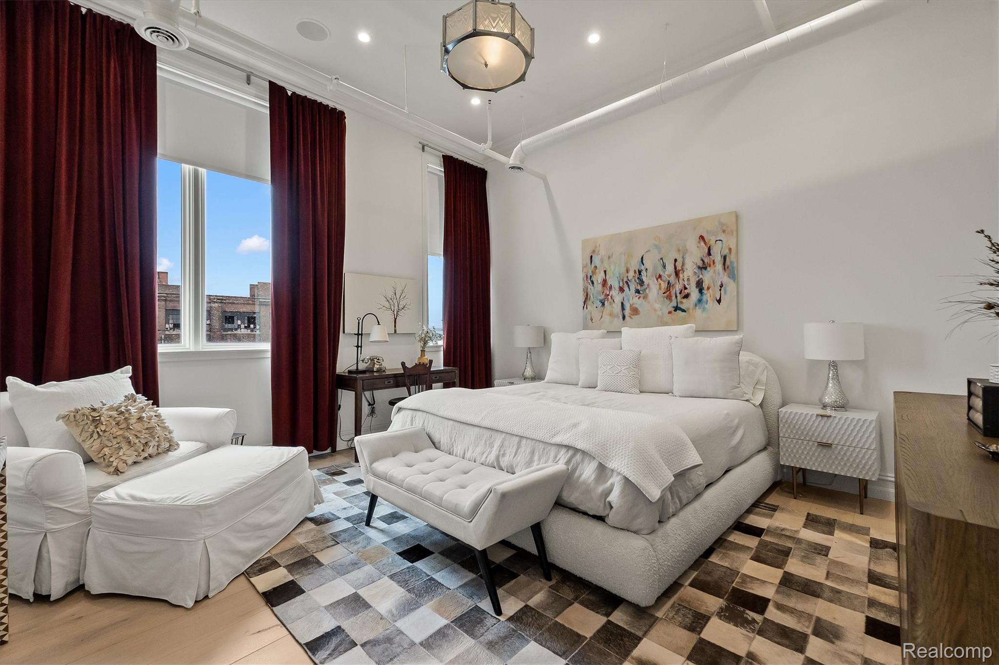 Bright bedroom with a large white bed, burgundy curtains, and a patchwork rug.