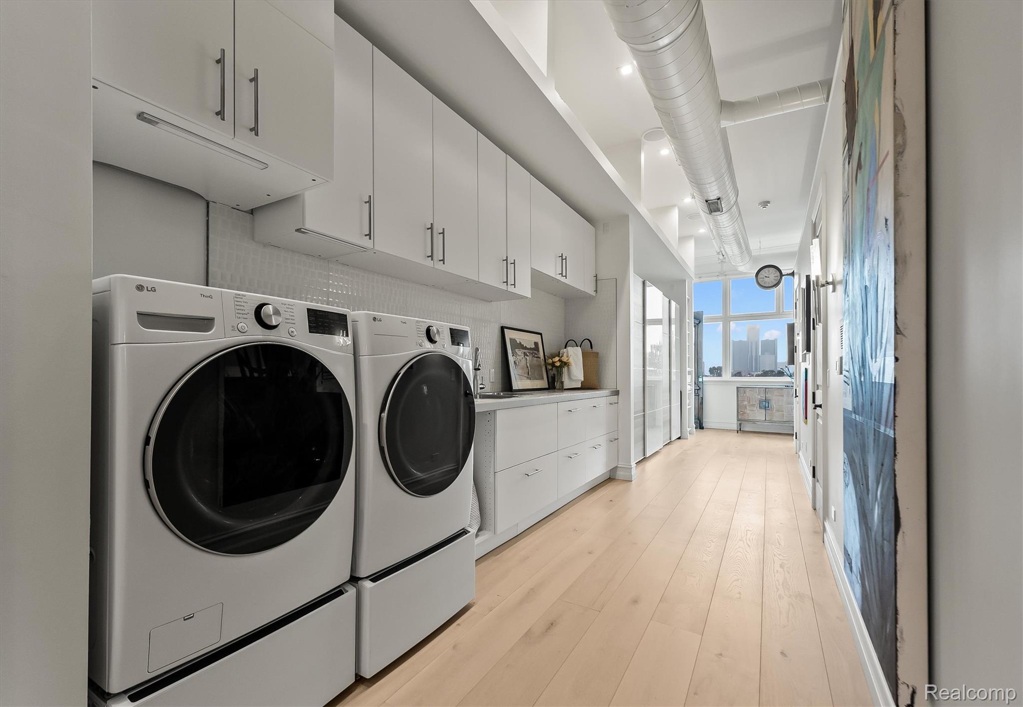 Modern laundry room with front-loading washer and dryer, white cabinetry, pale wood floors, and a Detroit skyline view.