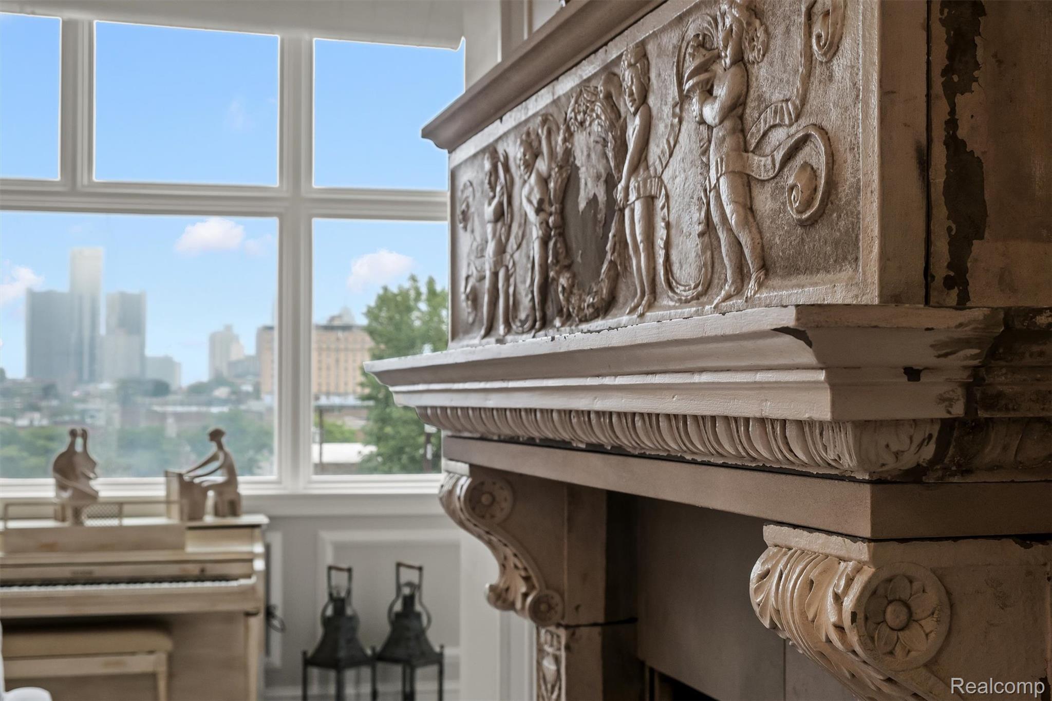 Ornate carved fireplace beside a piano, with tall windows overlooking the Detroit skyline.