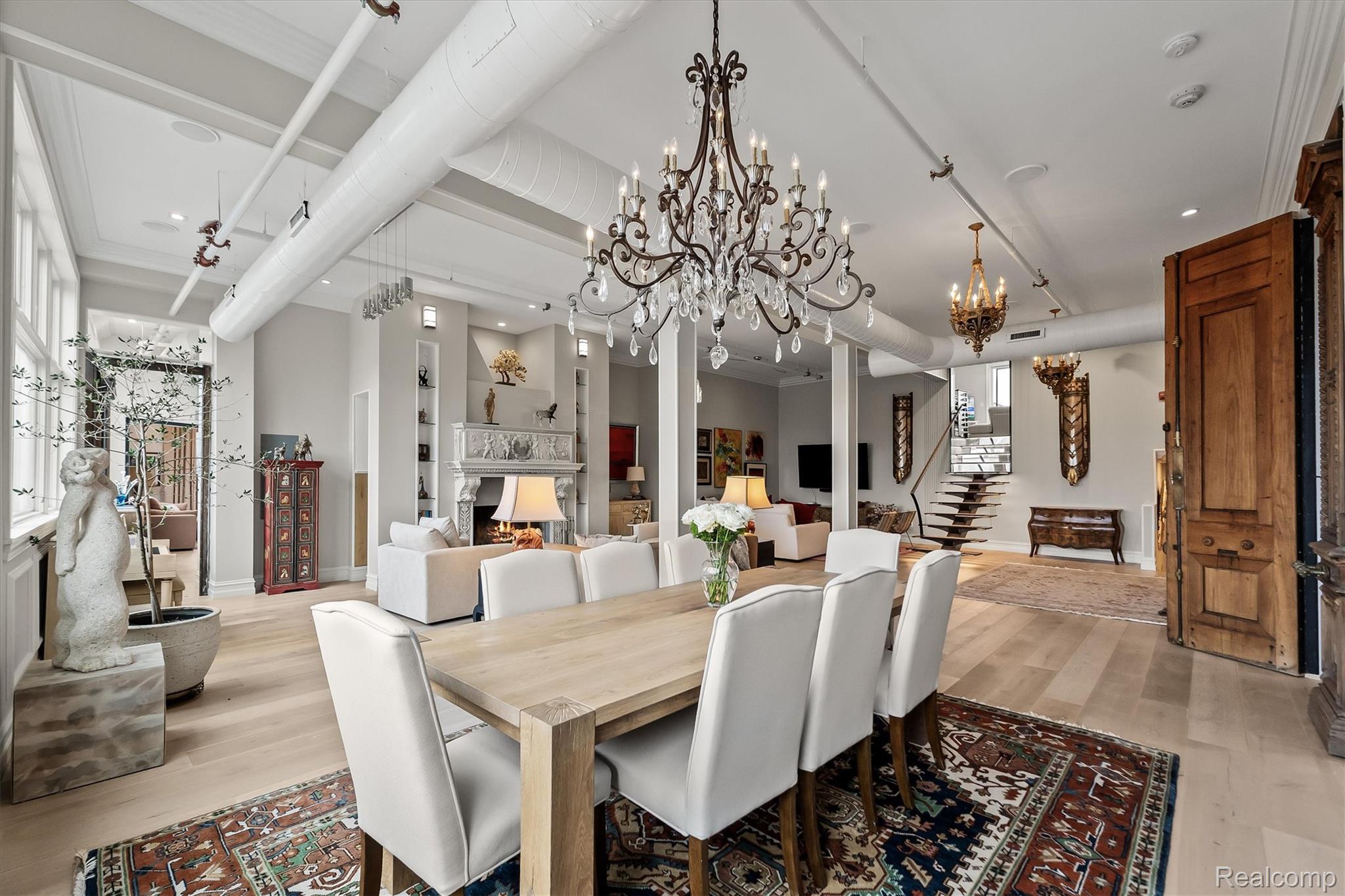 Open dining and living room with ornate chandeliers, a fireplace, exposed ducts, and a floating staircase.