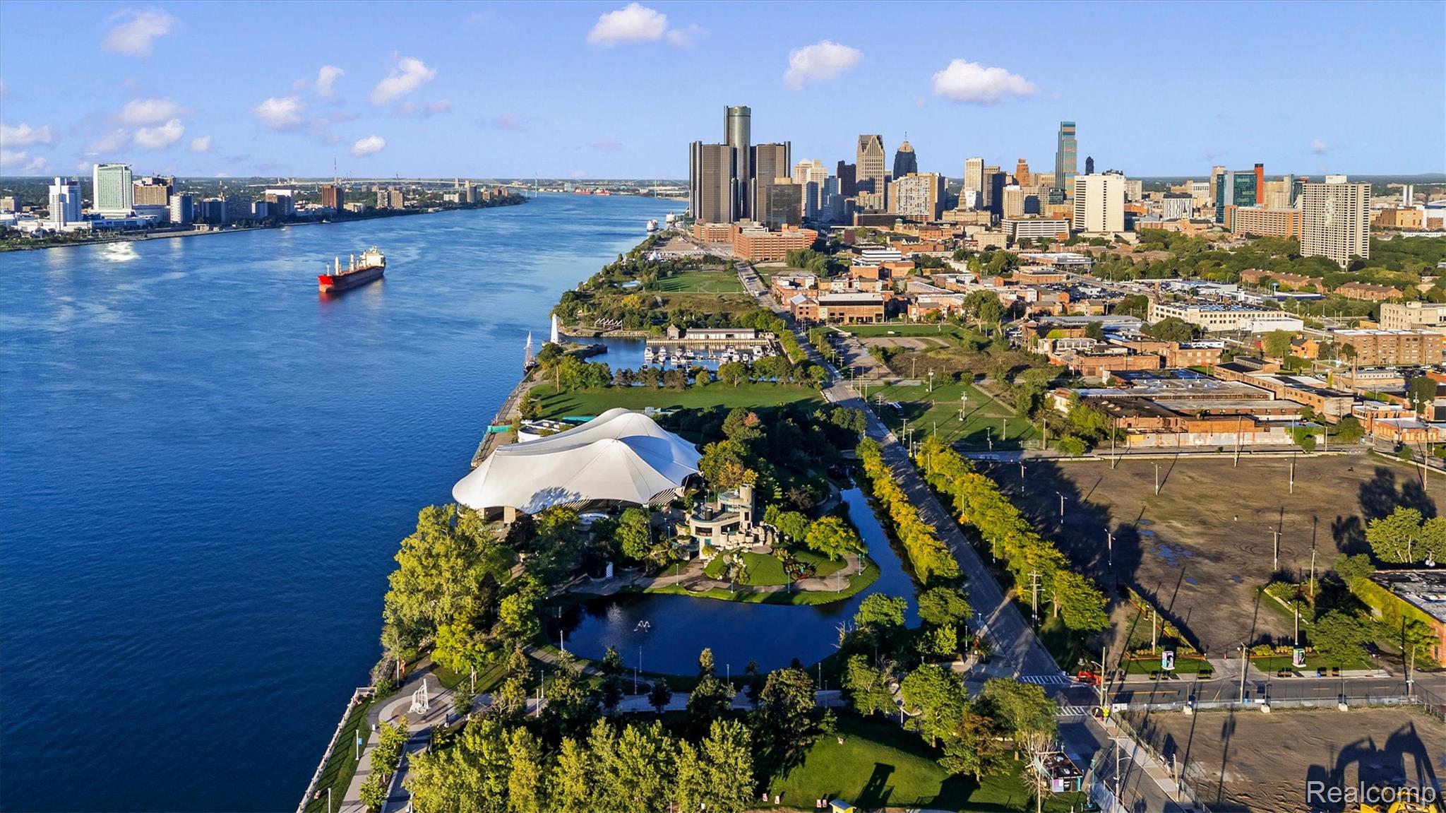 Aerial view of downtown Detroit and the river, with a freighter and a white-roofed pavilion.