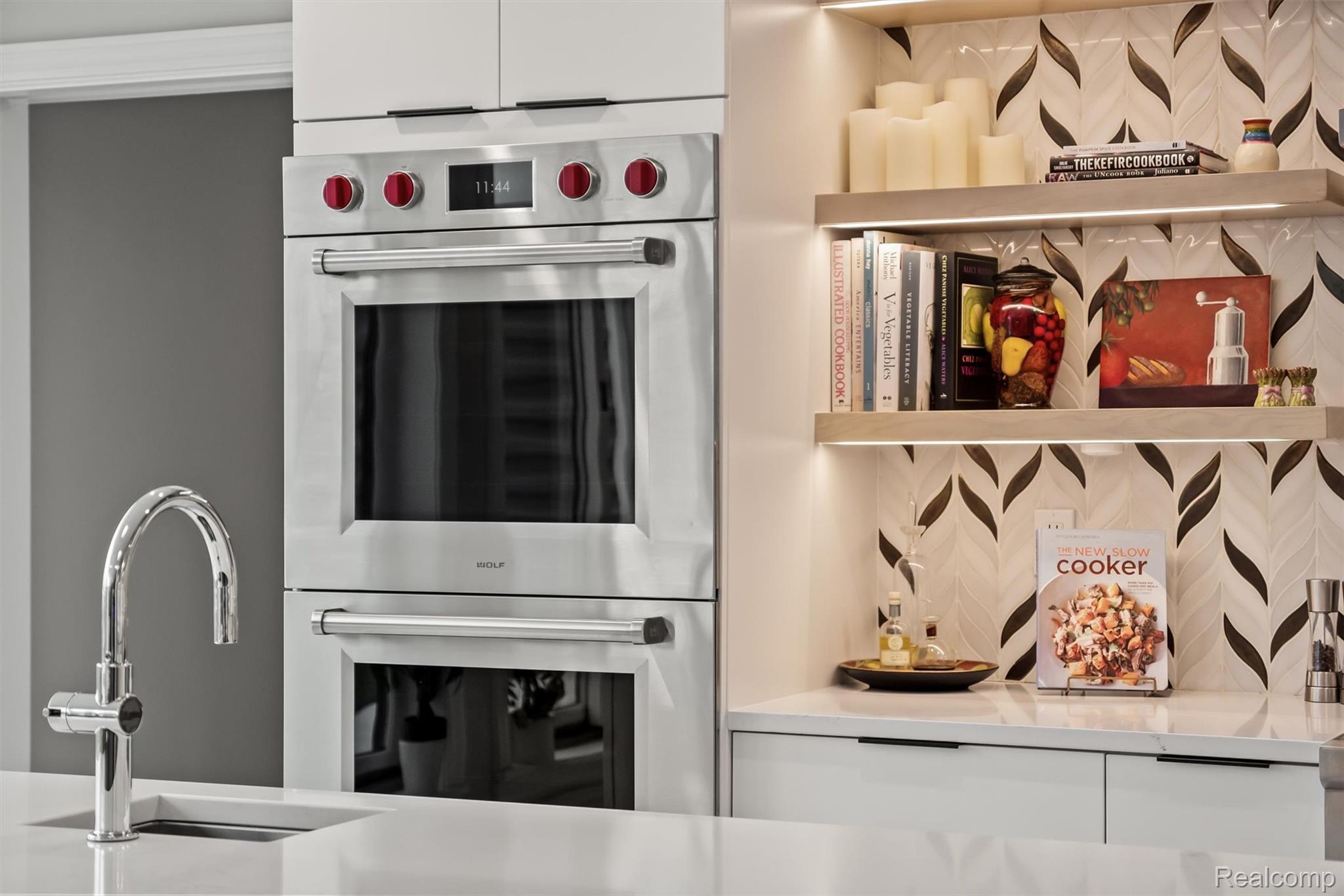 Modern kitchen with stacked stainless-steel ovens, red knobs, and illuminated cookbook shelves.