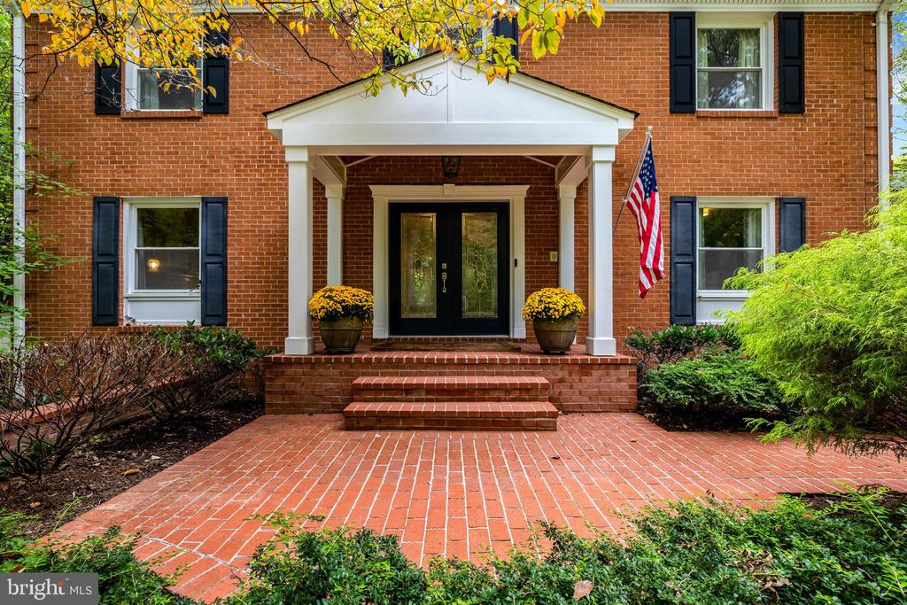 Red brick home entrance with a white portico, brick walkway, autumn leaves, and an American flag.