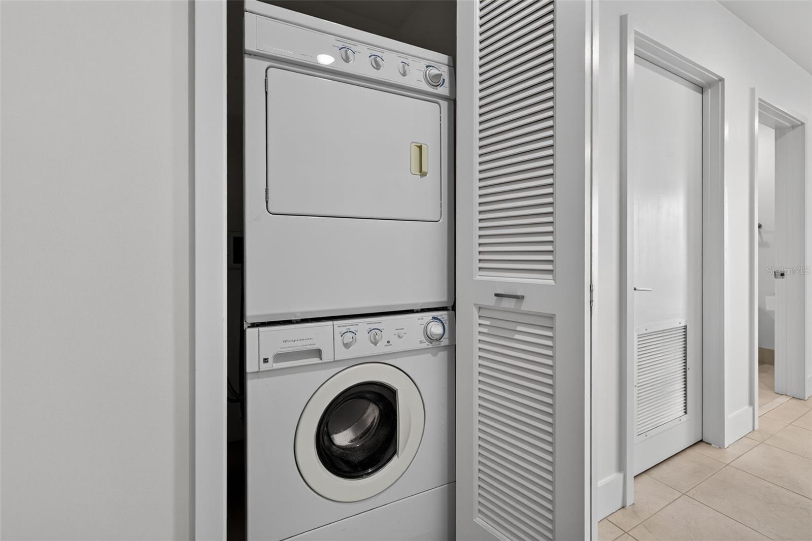 Stacked washer and dryer in a recessed laundry closet beside louvered doors.