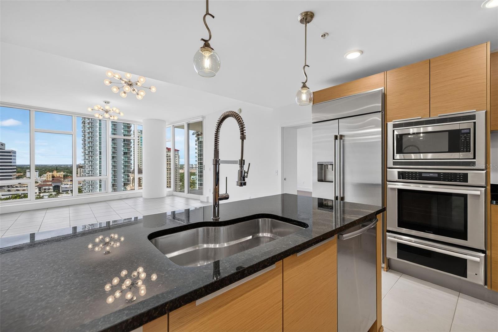 Modern condo kitchen with a dark stone island, stainless appliances, and high-rise views through floor-to-ceiling windows.