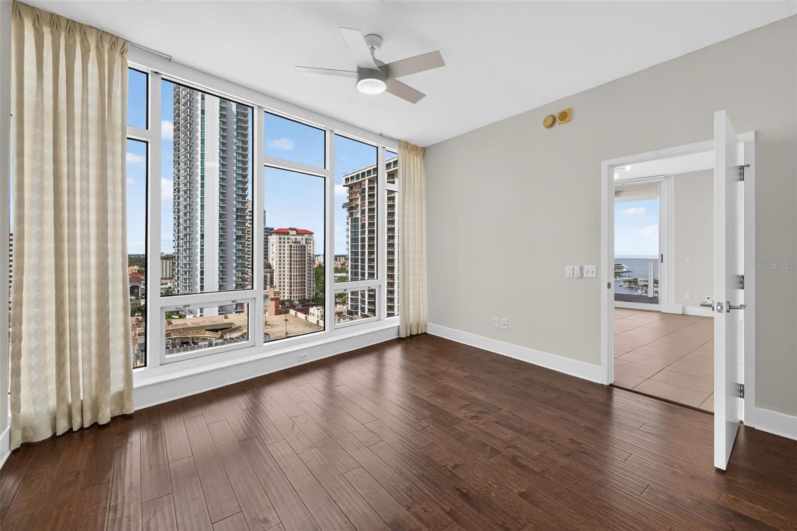 Empty condo room with dark wood floors, floor-to-ceiling windows, and downtown views.