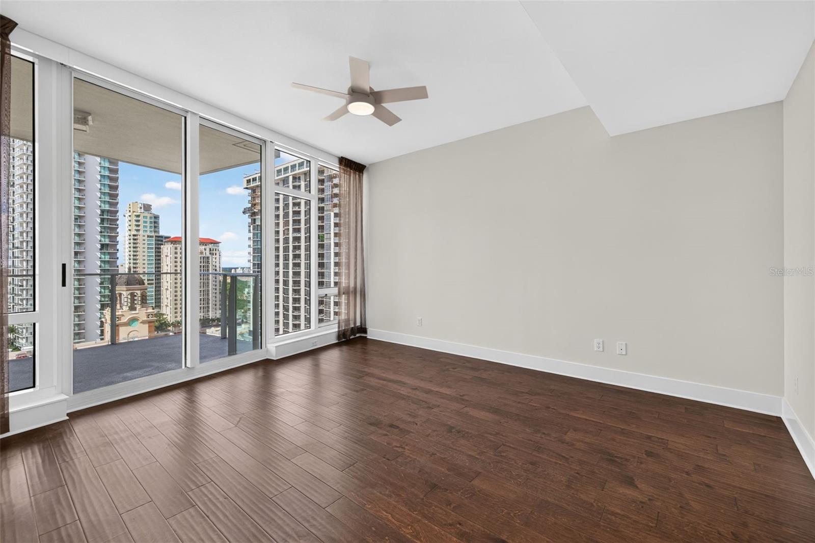 Empty condo living room with dark wood flooring and floor-to-ceiling windows overlooking city towers.