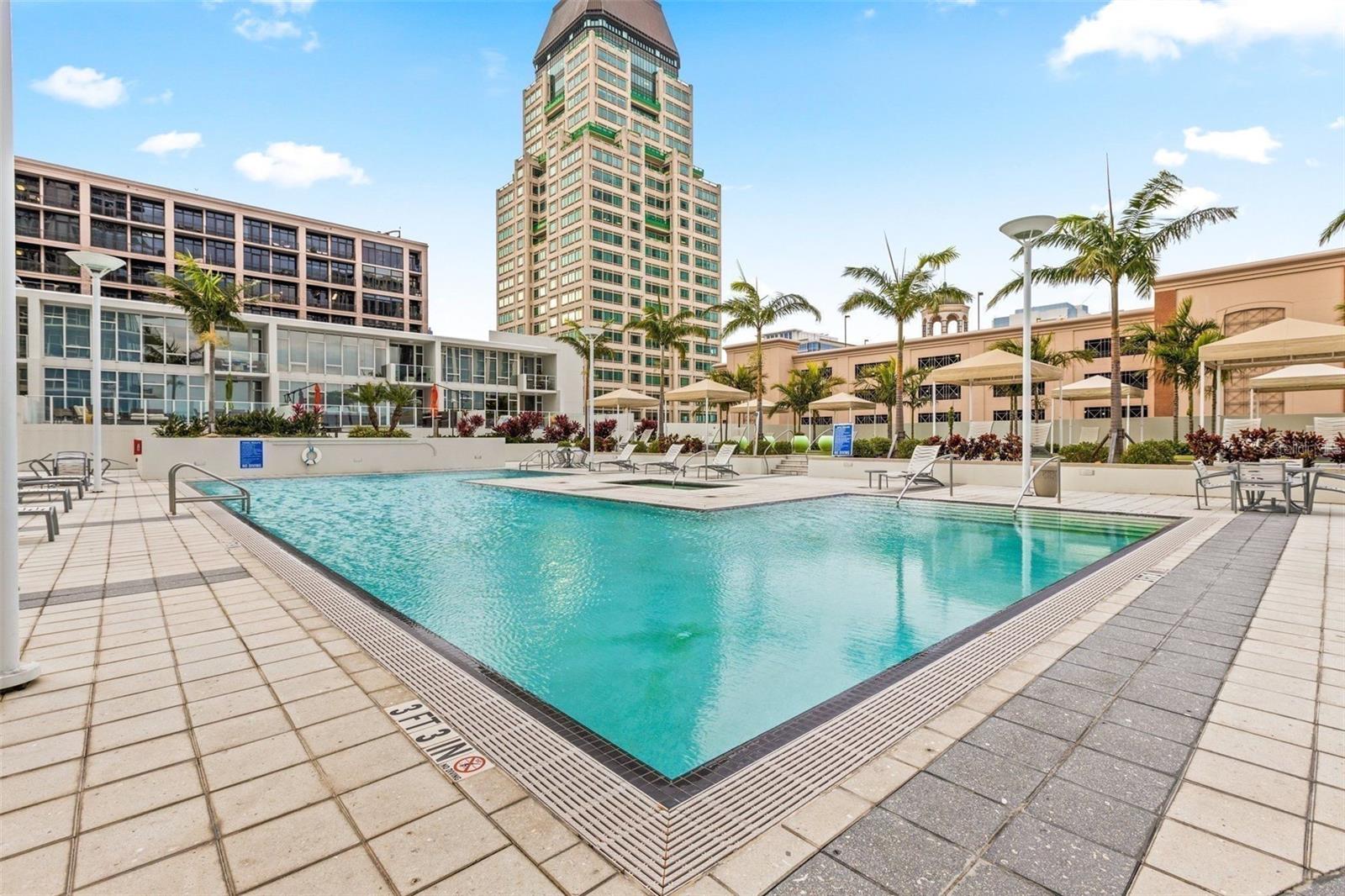 Outdoor condominium pool with a tiled deck, lounge chairs, umbrellas, and palm trees against downtown high-rises.