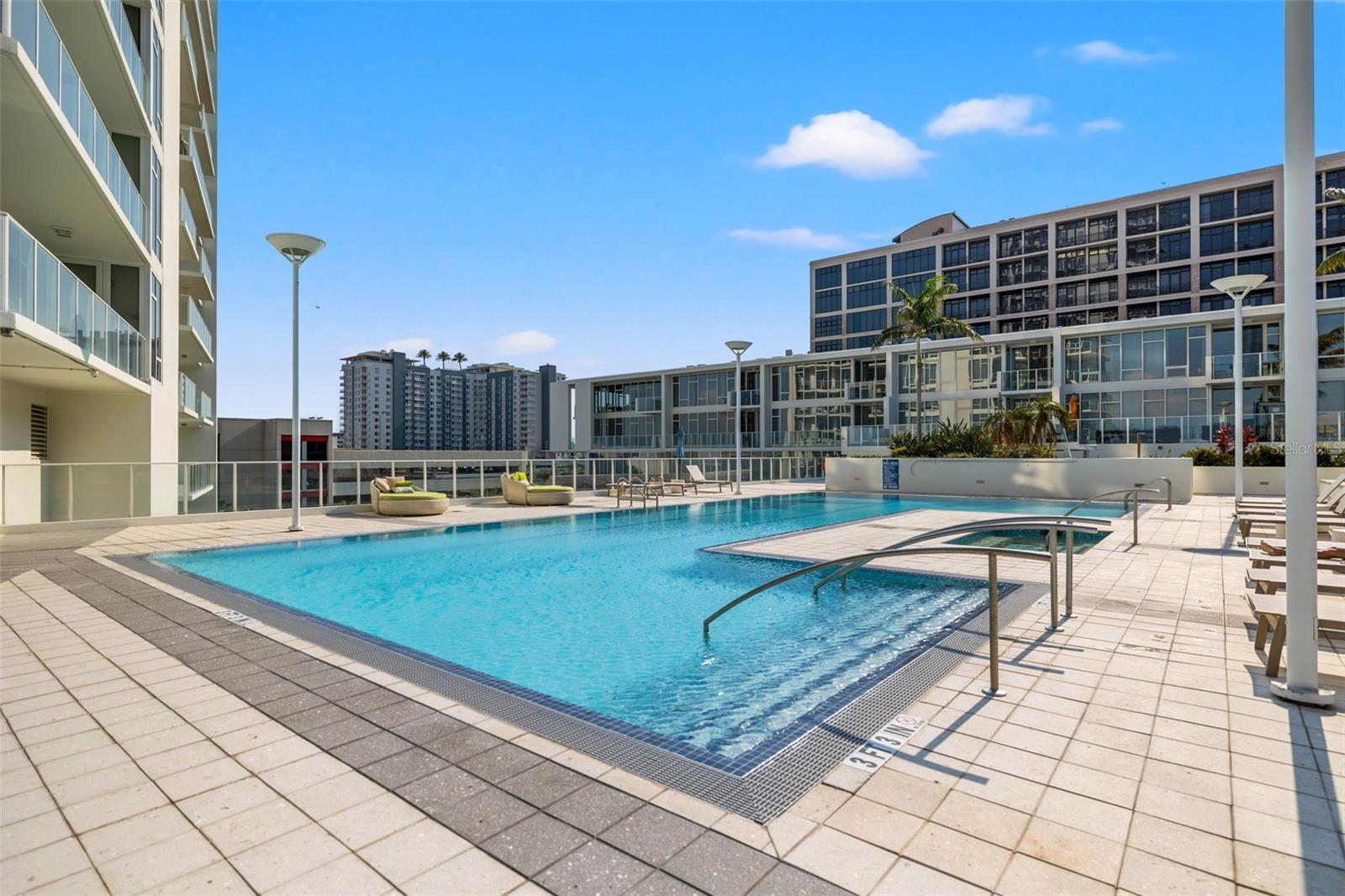 Condominium pool with tiled deck, raised spa, and lounge chairs surrounded by modern buildings.