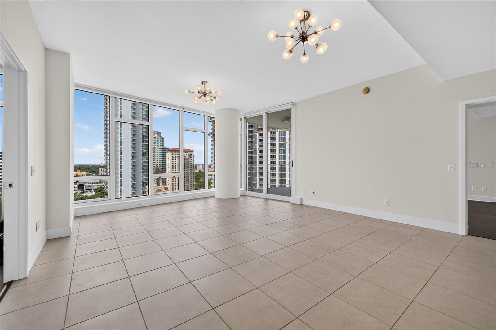 Vacant condo living room with tiled floors, modern chandeliers, and floor-to-ceiling windows overlooking city buildings.