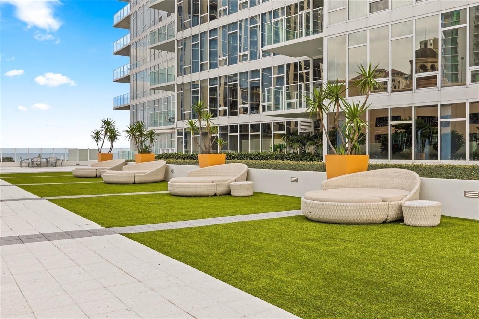 Condominium terrace with curved lounge seating, orange planters, green lawn, and a glimpse of water.