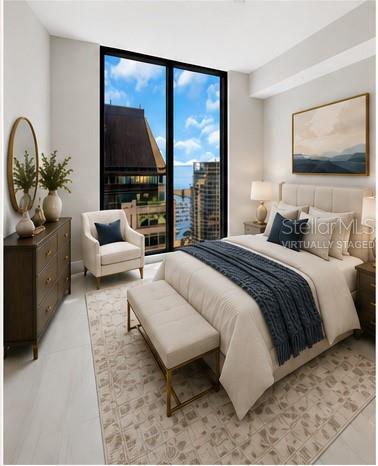 Staged bedroom with a king bed, upholstered bench, and floor-to-ceiling windows overlooking city buildings.