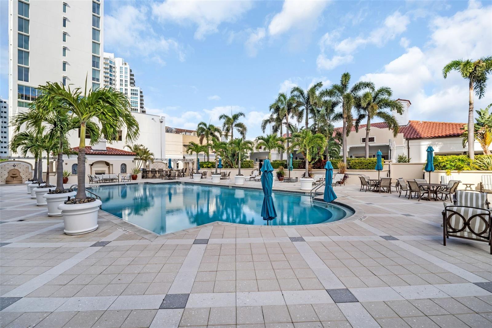 Condominium pool courtyard with palm trees, tiled deck, and blue umbrellas.