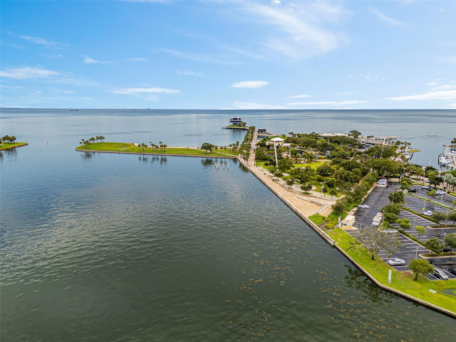 Aerial waterfront view with a landscaped promenade, palm-lined green space, docks, and calm open water.