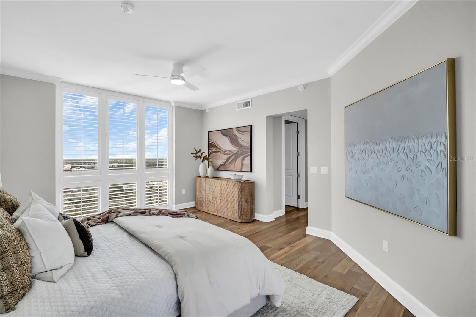 Bedroom with a white bed, wood floors, and large shuttered windows overlooking nearby buildings and greenery.