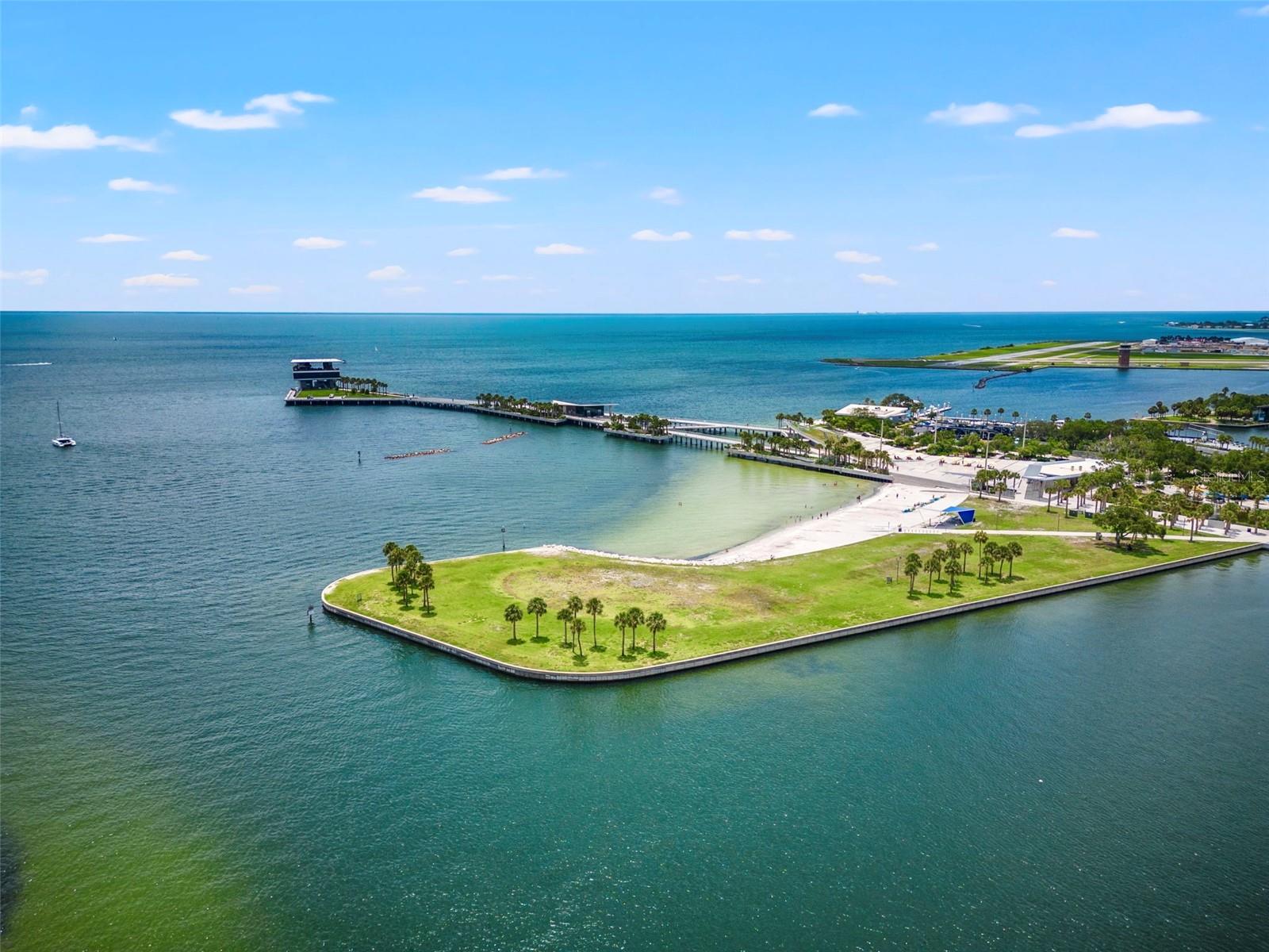 Aerial view of a palm-lined waterfront park, sandy beach, and long pier surrounded by blue water.