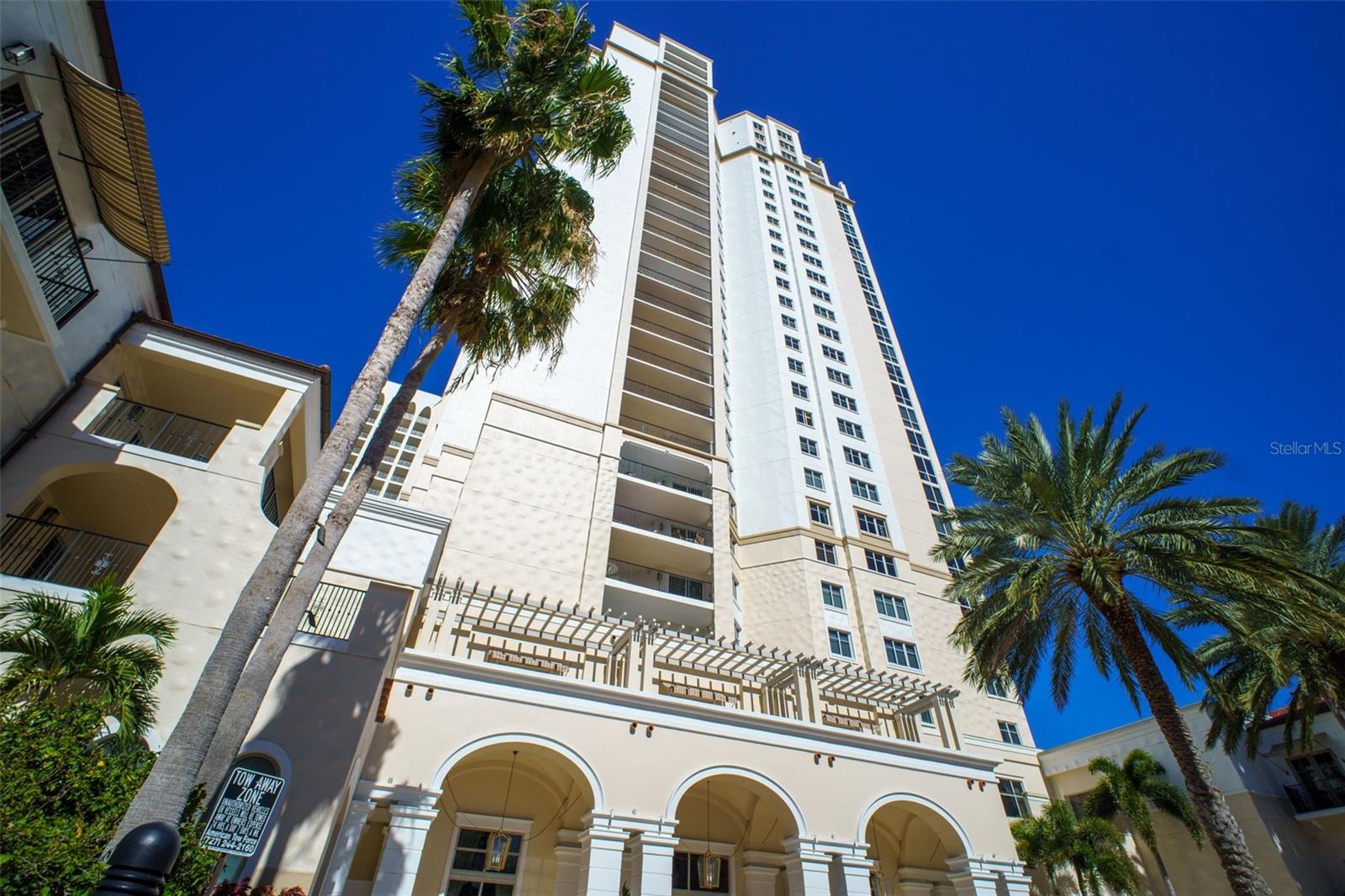 White condominium tower with balconies and arched entrances, framed by tall palm trees.