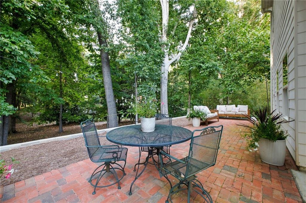 Brick patio with metal dining set and cushioned sofa beside a wooded yard.