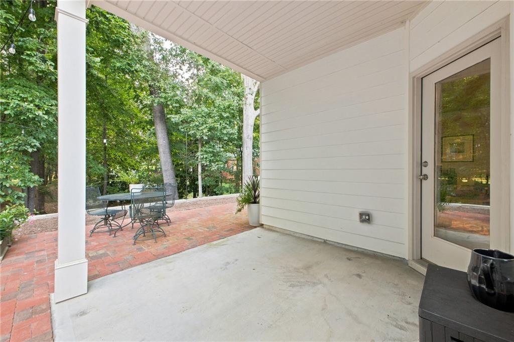 Covered patio opening onto a brick terrace with metal seating and a leafy wooded backdrop.