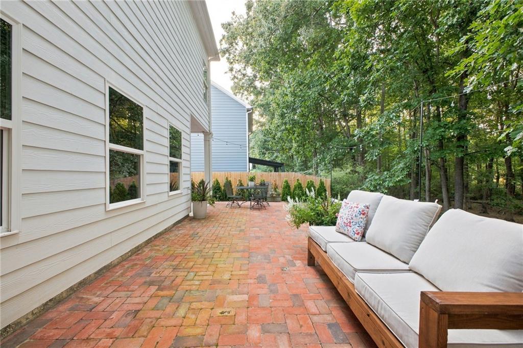 Brick patio beside a light-sided house, furnished with a white-cushioned sofa and surrounded by trees.
