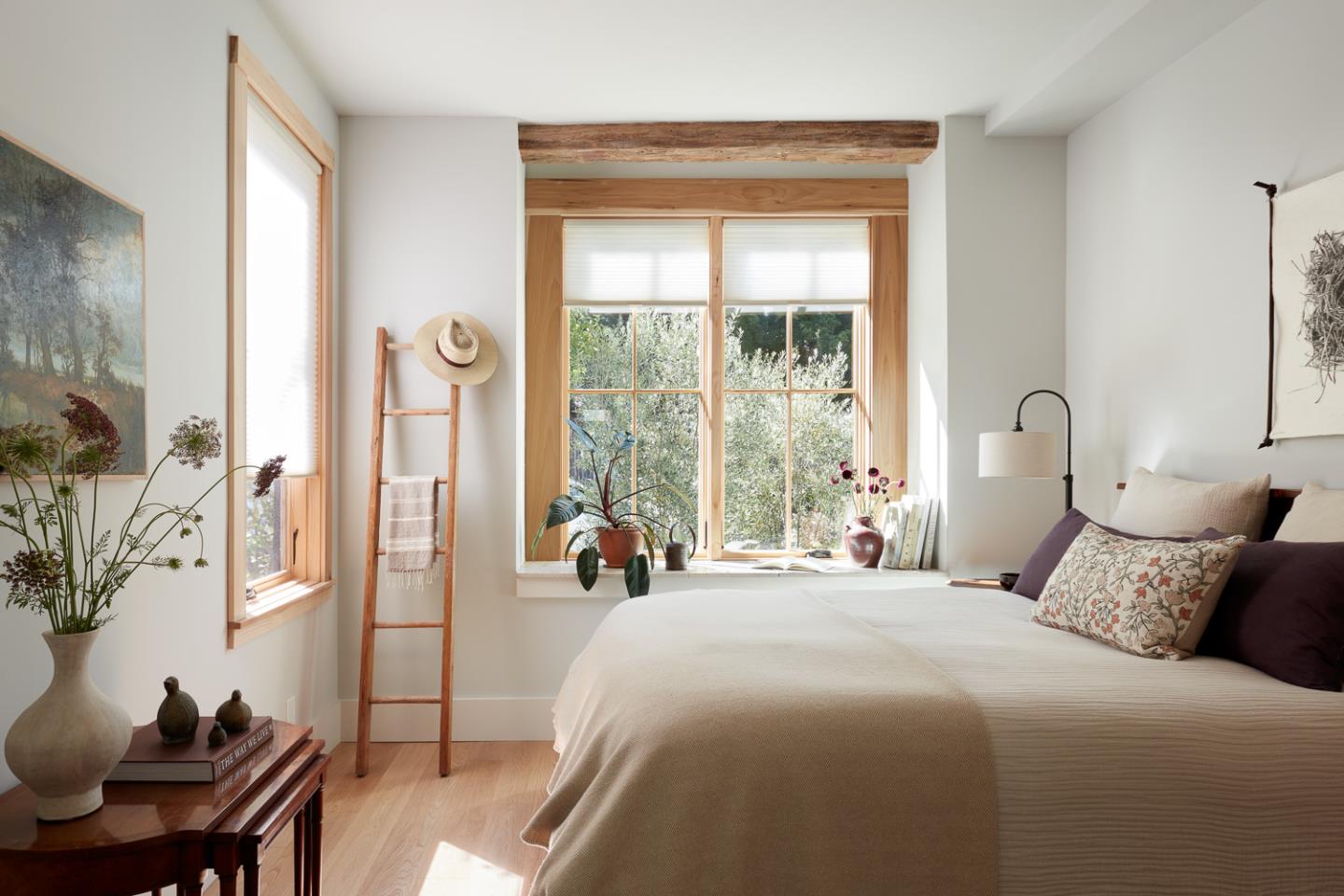 Sunlit bedroom with a large white bed, wood-framed windows, and a rustic beam.