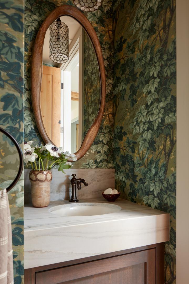 Powder room vanity with leafy wallpaper, an oval wood-framed mirror, and a white stone countertop.