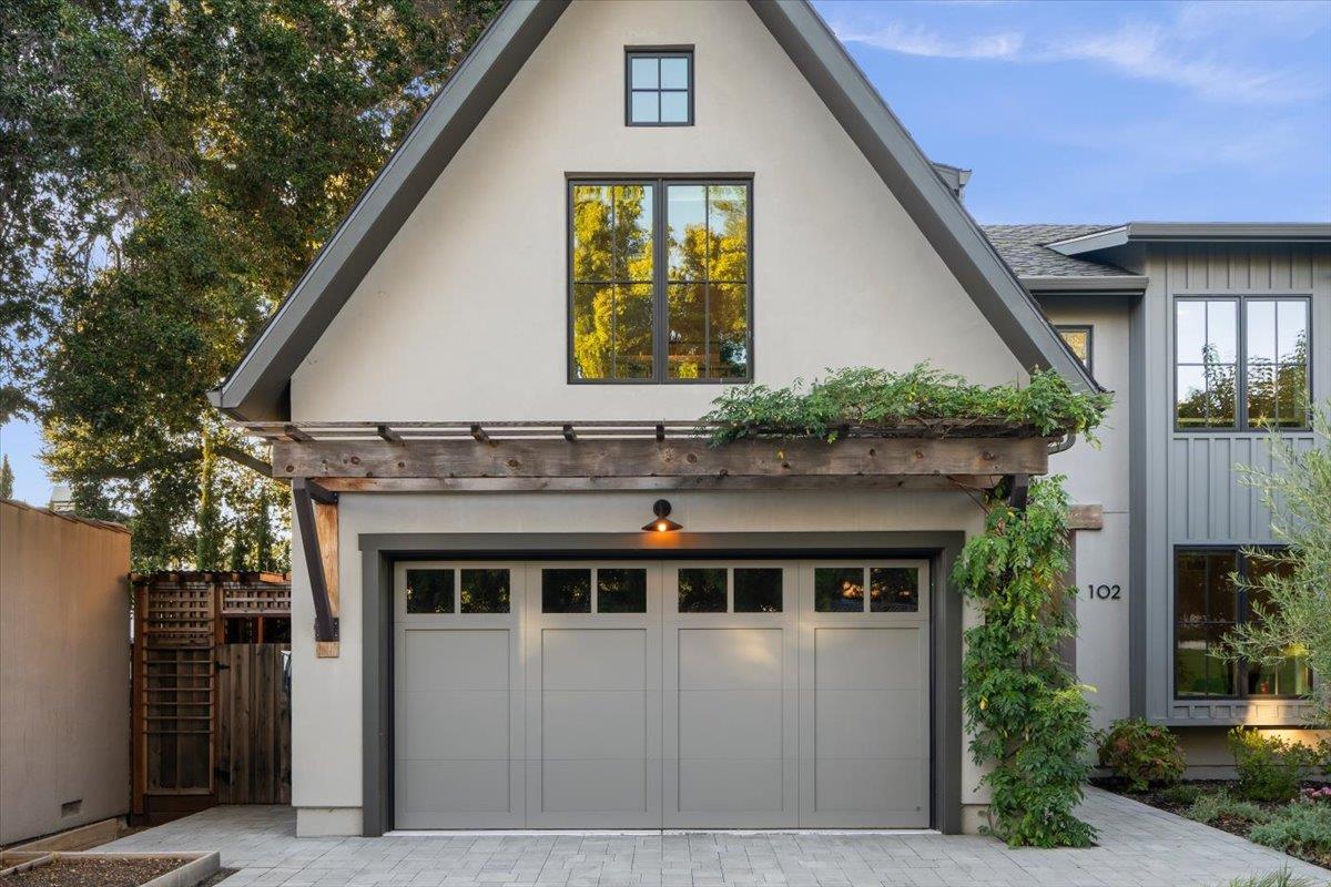 Home exterior with a steep gable, wide garage door, and greenery-covered pergola.