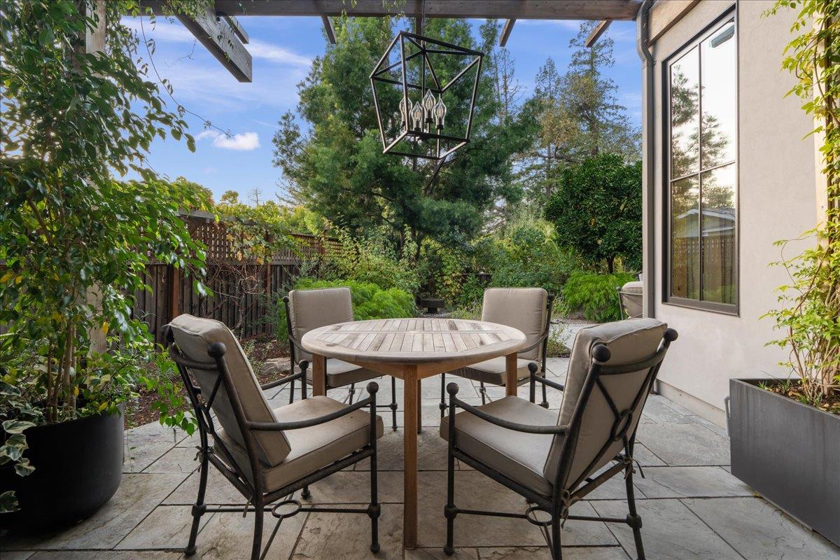 Outdoor patio with a round dining table, four cushioned chairs, stone pavers, and a leafy garden.