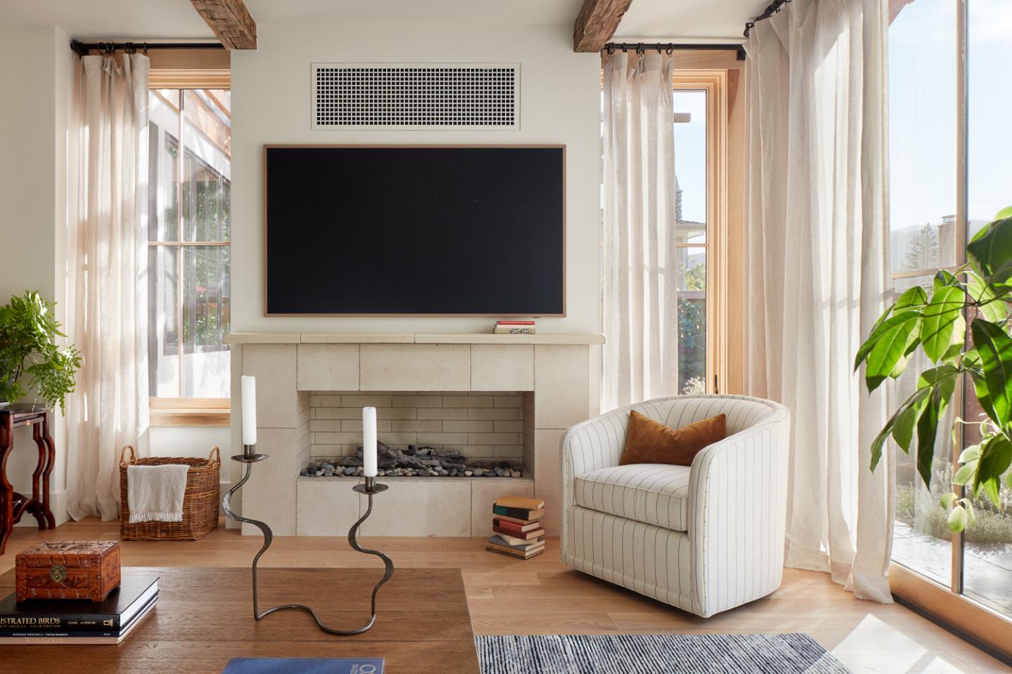 Living room with a stone fireplace, large screen, exposed beams, and a striped armchair beside tall windows.