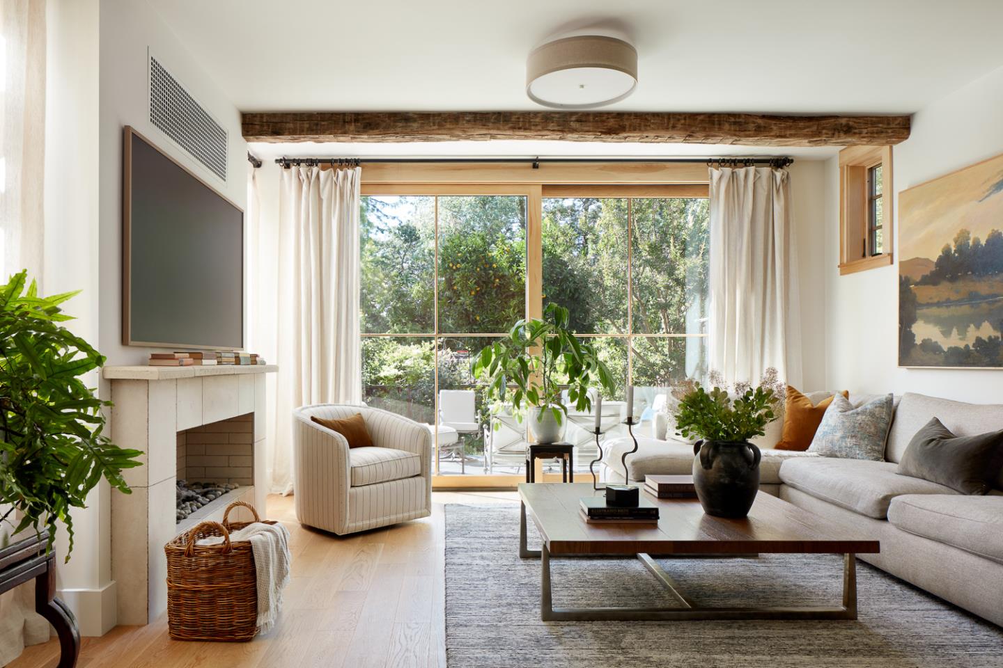 Living room with a stone fireplace, exposed beam, and large glass doors overlooking greenery.