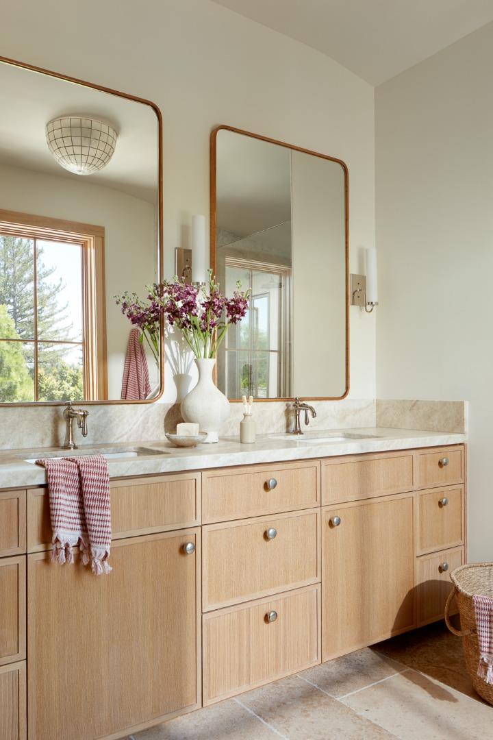 Bathroom vanity with pale wood cabinets, two framed mirrors, and flowers on the countertop.