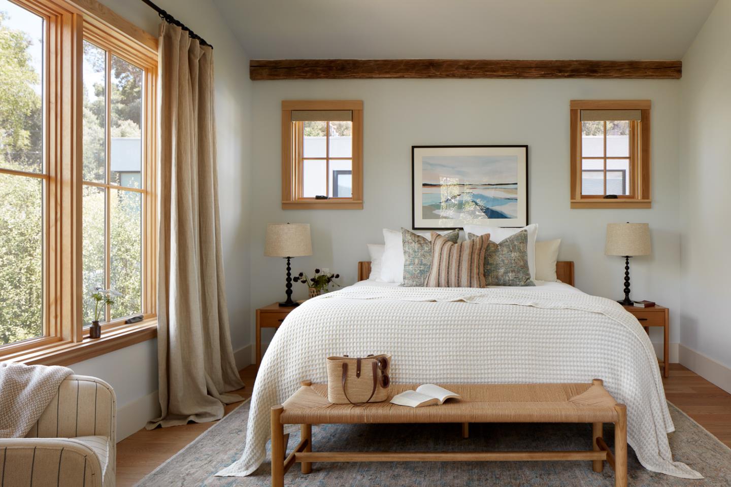 Staged bedroom with a white bed, rustic ceiling beam, and large windows overlooking trees.