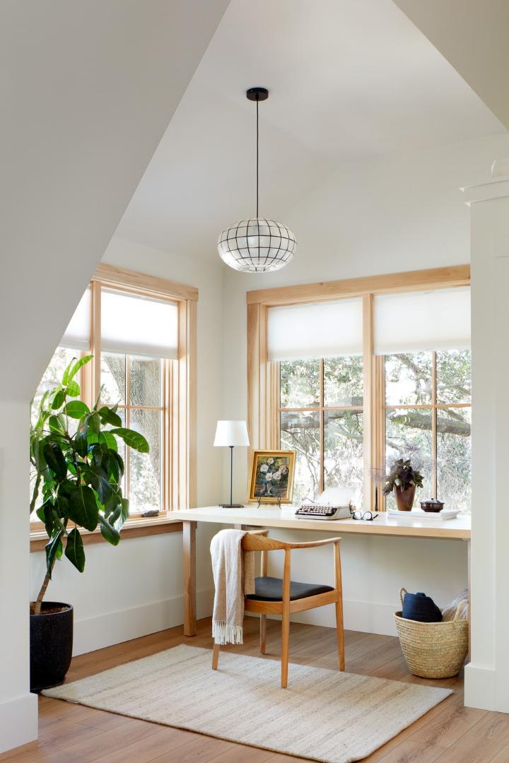 Bright home office with a desk beneath large windows, a wooden chair, and a potted plant.