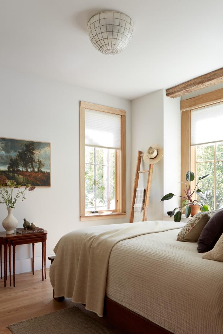 Bright bedroom with a large bed, tall wood-trimmed windows, and leafy plants.