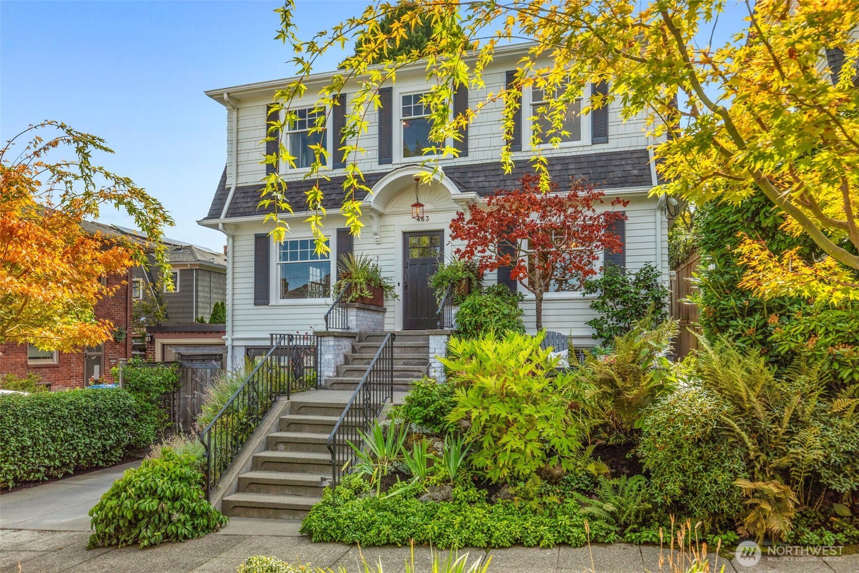 White two-story house with dark shutters, broad front steps, and colorful autumn trees and garden plantings.