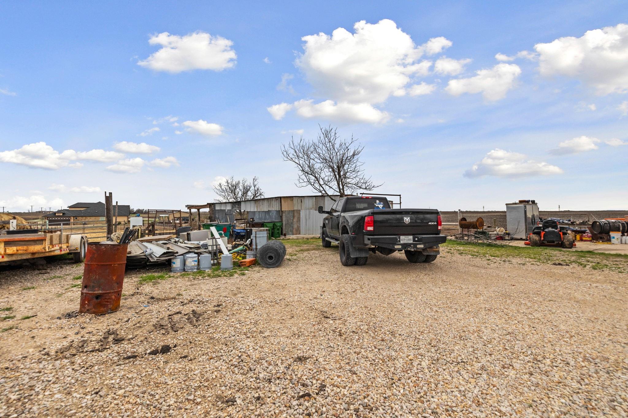 Gravel property yard with a black pickup, metal sheds, and scattered equipment and materials.