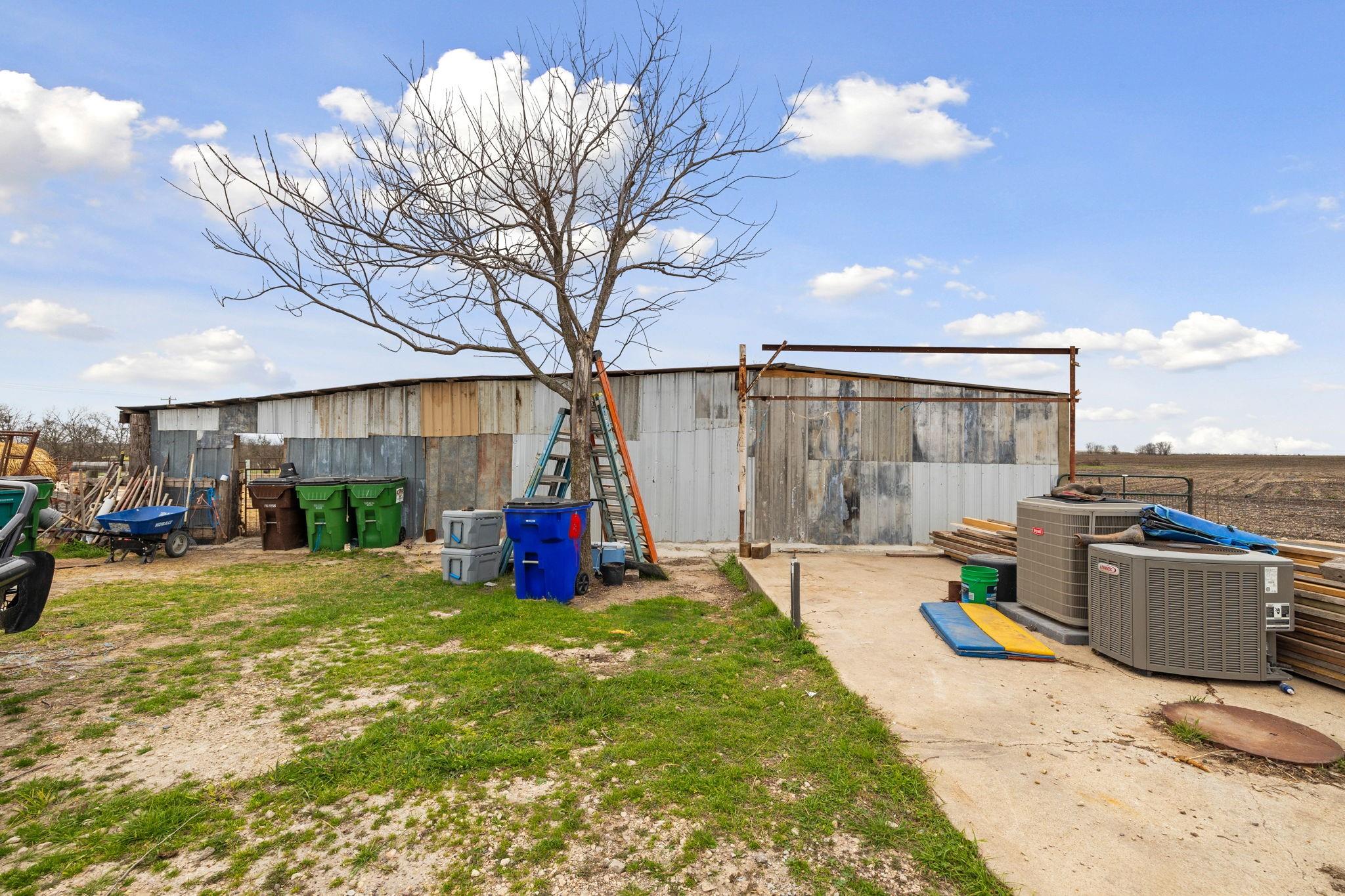 Weathered metal outbuilding beside a leafless tree, with ladders, bins, lumber, and air-conditioning units.