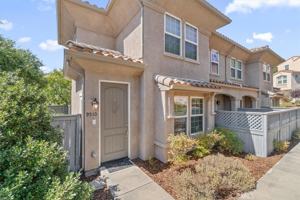 Two-story tan stucco townhouse with a tiled roof, gray fence, and 9510 beside the front door.
