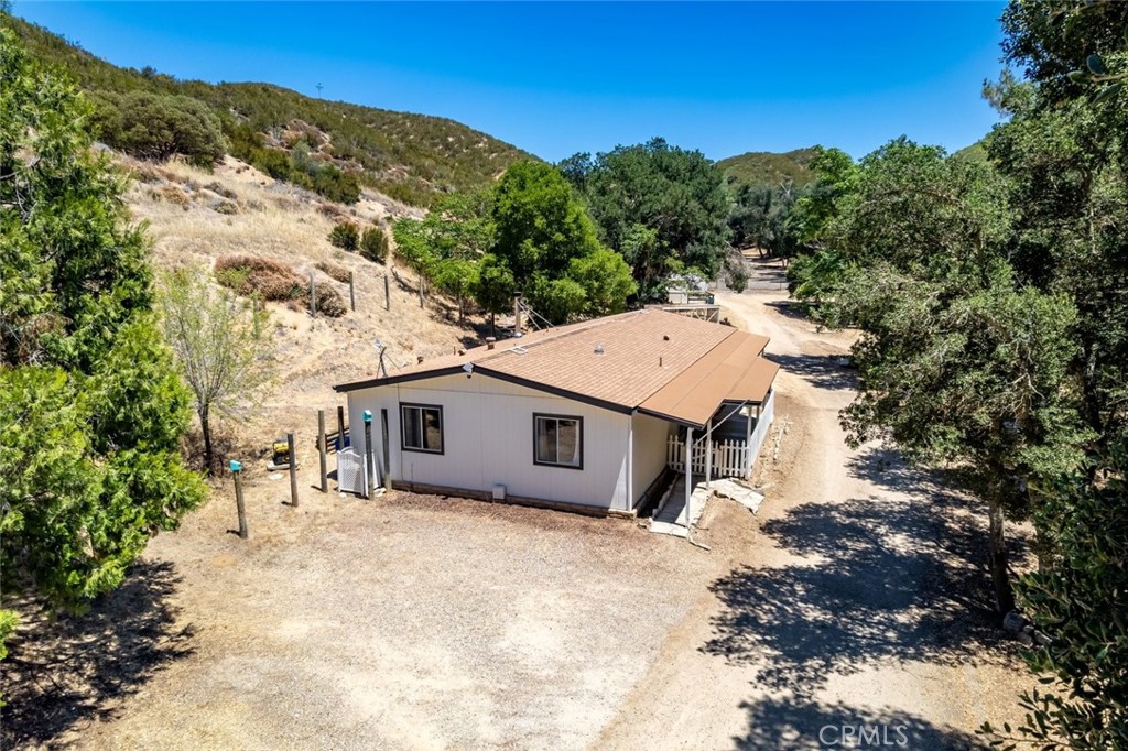 Aerial view of a single-story home with a tan roof beside a dirt driveway and dry hillsides.