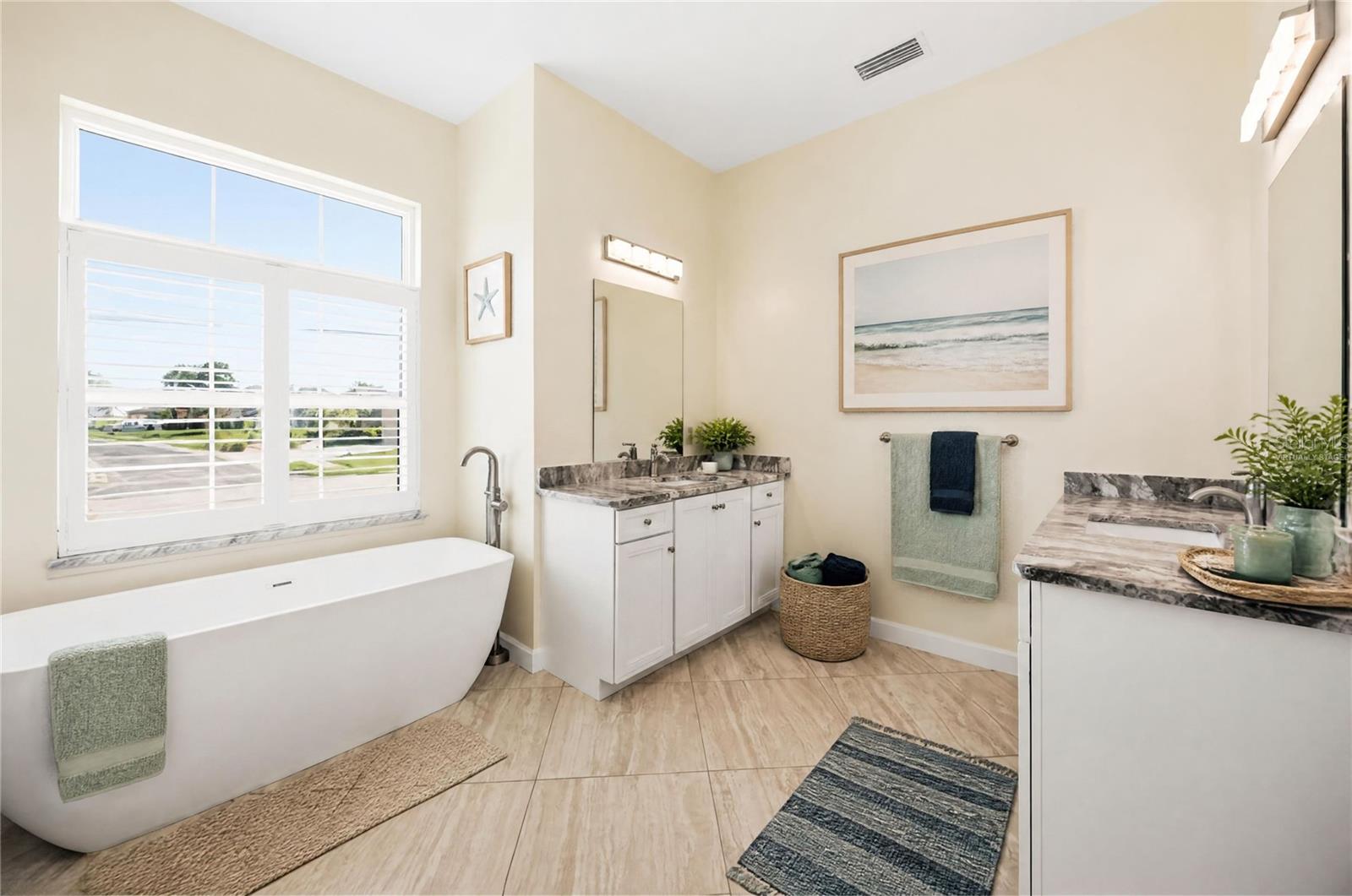 Bathroom with a freestanding tub under a large window and two marble-look vanities.