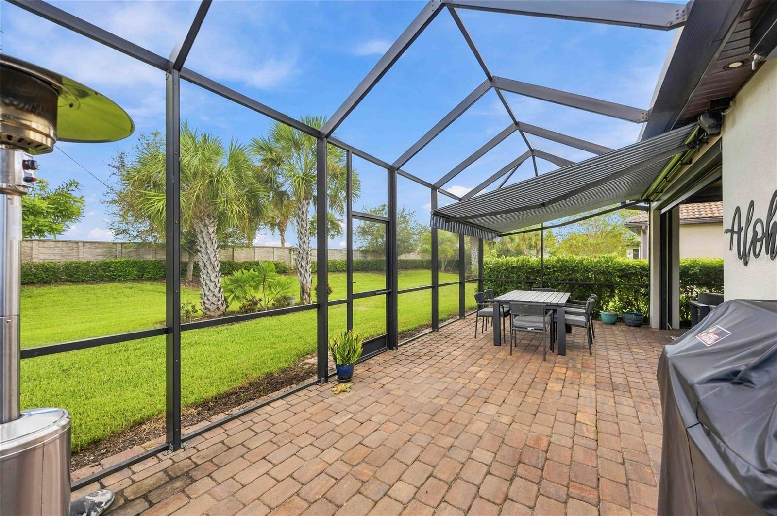 Screened lanai with a brick patio, retractable awning, and outdoor dining area overlooking a palm-lined lawn.