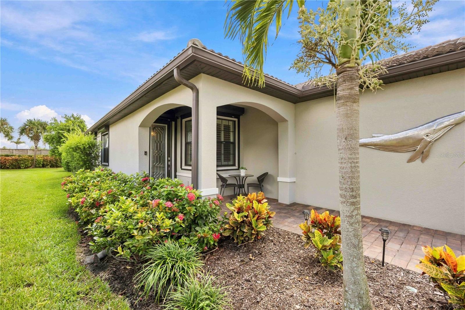Stucco home exterior with arched covered entry, patio seating, tropical landscaping, and a fish wall sculpture.