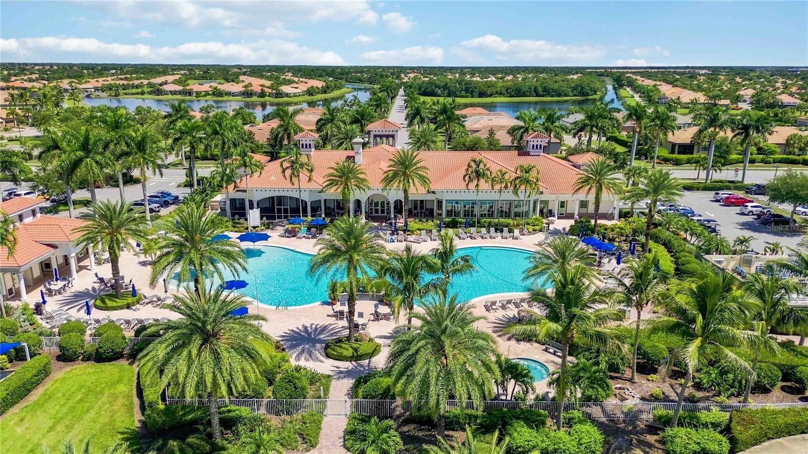 Aerial view of a community clubhouse and two pools surrounded by palm trees, with lakes in the distance.